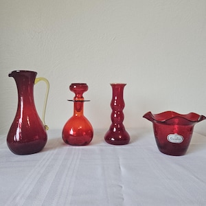 May include: Four pieces of red glass home decor. Includes a pitcher with a yellow handle, a bottle with a stopper, a tall vase, and a ruffled bowl. The items are arranged on a white tablecloth.