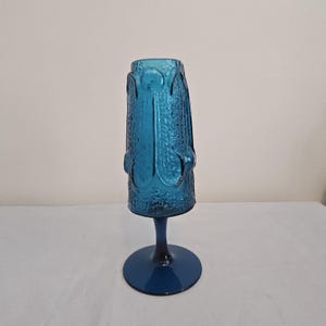May include: A tall, blue glass vase with a textured surface and a pedestal base. The vase has a cylindrical shape with decorative vertical and circular patterns. The base is round and the vase is approximately 10 inches tall.