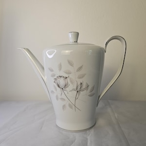 May include: White porcelain coffee pot with a lid and handle. The pot features a floral design of a rose and leaves in gray tones. Silver trim accents the edges, handle, and lid.