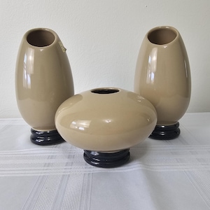 May include: Set of three ceramic vases in a light brown color with a glossy finish. The vases have a rounded shape and a dark blue base.
