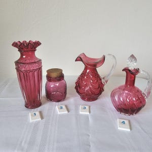 May include: A collection of four antique cranberry glass pieces. Includes a tall vase with a ruffled rim, a small jar with a cork stopper, a pitcher with a clear handle, and a decanter with a glass stopper and handle.