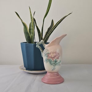 May include: A vintage pink ceramic pitcher with a floral design and a pedestal base. The pitcher has a curved spout and a handle with a light blue accent. A blue hexagonal planter with a snake plant is in the background.
