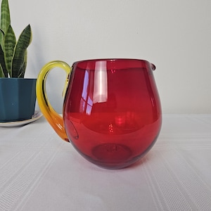 May include: A red glass pitcher with a yellow handle. The pitcher has a rounded body and a wide mouth.