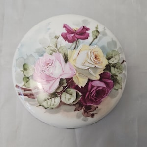 May include: A round, white ceramic trinket box with a hand-painted floral design. The lid features a bouquet of roses in shades of pink, yellow, and deep purple, surrounded by green leaves. The box has a glossy finish.