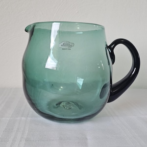 May include: A translucent seafoam green glass pitcher with a rounded body, a small pouring spout, and a dark black glass handle. The front of the pitcher features a small sticker with the text Blenko Handcraft Made in USA printed on it.
