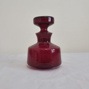 May include: A ruby red glass decanter with a matching stopper. The decanter has a rounded base, a slender neck, and a flared top. The stopper is a flat disc shape. The glass has a glossy finish.
