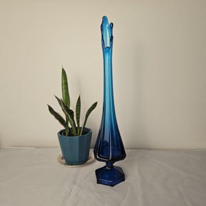May include: A tall, vintage blue glass vase with a flared top and a geometric base. The vase is approximately 24 inches tall. A potted snake plant in a blue pot sits to the left of the vase.