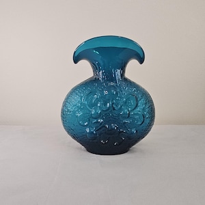 May include: A teal-colored glass vase with a textured surface and a unique, flared top. The vase has a round body with embossed floral patterns. The overall design is a blend of vintage and modern aesthetics, suitable for home decor.