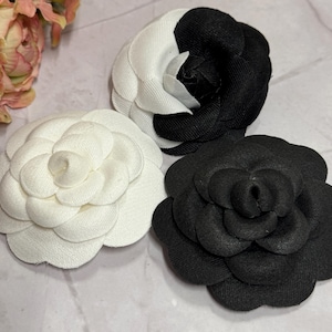May include: Three fabric flower brooches in a close-up shot. One is solid white, another is half black and half white, and the third is solid black. The brooches are in the shape of camellias, with layered petals.