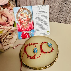 Puede incluir: Dos pulseras de cuentas rojas con pequeños colgantes ovalados con una imagen religiosa, apoyadas sobre una superficie crema con bordes dorados. Una tarjeta con texto en español y una imagen religiosa está detrás de las pulseras. Las pulseras son un artículo religioso.