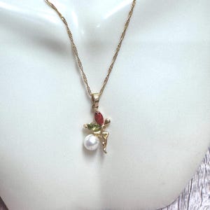 May include: A gold-tone necklace featuring a pendant shaped like a figure with a white spherical bead, a marquise-cut red stone, and a marquise-cut green stone. The pendant hangs from a delicate gold-tone twisted chain on a white display bust.