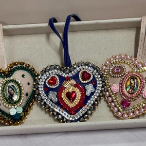 Peut inclure: Trois ornements en forme de cœur avec des perles complexes et des images religieuses. Les ornements présentent diverses couleurs, dont le rose, le bleu et l'or, et sont exposés dans un plateau rectangulaire. Chaque cœur a une boucle pour accrocher.
