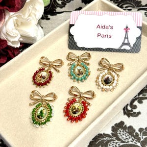 May include: A collection of ornate brooches with gold-tone bows and oval pendants. Each brooch features a central image surrounded by colorful beads in red, green, turquoise, and white. The brooches are displayed in a cream-colored tray.