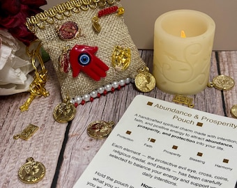 May include: A beige burlap pouch with gold trim, adorned with charms including a red hand, gold coins, and a cross. A lit candle and a card with text about abundance and prosperity are also present.