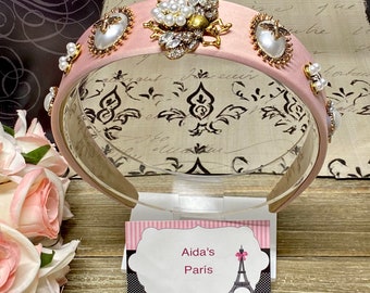 May include: Pink satin headband with gold and rhinestone embellishments, including a bee design with pearl accents. The headband is displayed on a stand with a sign that reads "Aida's Paris".