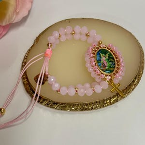 May include: Pink beaded bracelet featuring a gold cross charm and a central oval pendant with an angel illustration. The bracelet has a pink cord closure and gold accents. The beads are a light pink color.