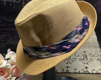 May include: A tan fedora hat with a plaid band in navy, pink, and gray. The hat is displayed on a black mannequin head. The hat has a woven texture and a classic fedora shape.