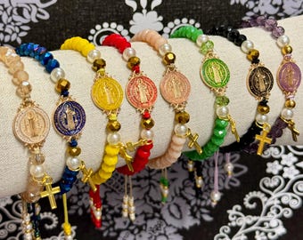 Puede incluir: Un conjunto de diez pulseras de cuentas de colores con dijes dorados que representan un icono religioso. Cada pulsera tiene un color de cuentas diferente y un dije de color diferente.