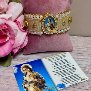 May include: A beige fabric bracelet featuring a central oval medallion with a religious image, surrounded by clear rhinestones and gold-tone accents. The bracelet is adorned with rows of small white beads and is accompanied by a prayer card with text.