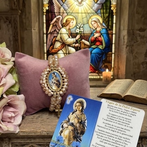 May include: A beaded rosary bracelet featuring a central oval medallion with a religious image, surrounded by light-colored beads and a small gold-toned cross charm. A prayer card with Spanish text and a religious illustration is placed in front.