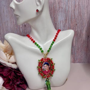Könnte beinhalten: Halskette mit einem Anhänger mit einem Porträt von Frida Kahlo, verziert mit roten und grünen Perlen. Der Anhänger ist von roten Perlen und goldfarbenem Metall umgeben. Eine grüne Quaste hängt darunter. Passende rote Ohrstecker sind ebenfalls sichtbar.