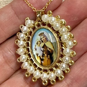 Puede incluir: Collar de tono dorado con un colgante ovalado de oro y perlas blancas que presenta una imagen religiosa de la Virgen María y el niño Jesús.