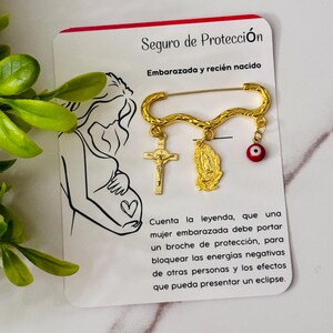 May include: A gold-colored safety pin with a crucifix, a Virgin Mary pendant, and a red evil eye charm. The pin is on a white card with text in Spanish, including "Seguro de Protección" and an illustration of a pregnant person.
