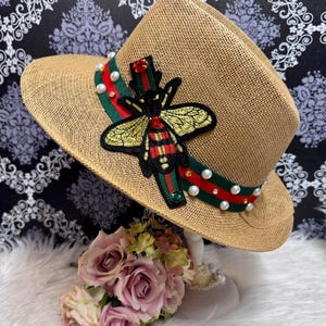 Puede incluir: Un sombrero fedora de paja color canela con una cinta a rayas verdes y rojas. El sombrero está decorado con un gran parche bordado de abeja con alas doradas, detalles corporales en rojo y negro, y pequeñas perlas blancas y cristales.