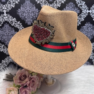 May include: A tan straw fedora hat featuring a wide green and red striped ribbon band. The hat is adorned with a large, three-dimensional heart-shaped patch covered in red crystals and gold-toned beadwork, accented by a single clear rhinestone.