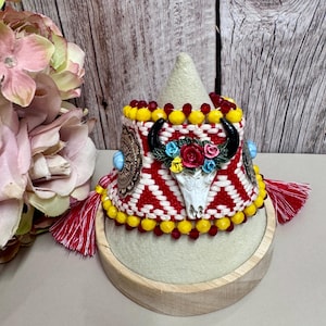 May include: A woven cuff bracelet featuring a red and white geometric pattern, accented with yellow and red beads. The center displays a white bull skull with colorful floral details, flanked by turquoise-colored stones and red tassels.