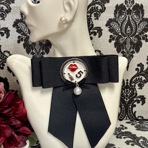 May include: A black ribbon bow tie with a circular brooch featuring red lips, a lipstick tube, and the number 5. A faux pearl hangs from the brooch. The bow tie is displayed on a white mannequin head with a pearl earring.