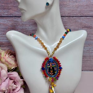 Könnte beinhalten: Eine farbenfrohe Perlenkette mit einem Anhänger, der Frida Kahlo zeigt. Der Anhänger ist von roten, blauen und goldenen Perlen umgeben, mit einer Quaste und einer kleinen blauen Blume. Ein passender blauer Ohrstecker ist ebenfalls sichtbar.