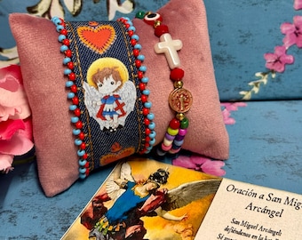 May include: Two bracelets with religious themes. One bracelet is made of blue fabric with red and white beads and features an embroidered image of an angel. The other bracelet is made of red and white beads with a white cross charm and a gold coin charm. The bracelets are displayed on a pink velvet cushion.