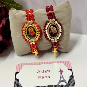 Puede incluir: Dos pulseras rojas con cuentas e imágenes religiosas, detalles dorados y pequeñas cruces doradas. Las pulseras presentan piezas centrales ovaladas con ilustraciones detalladas. Una tarjeta de visita con el texto "Aida's París" está en primer plano.