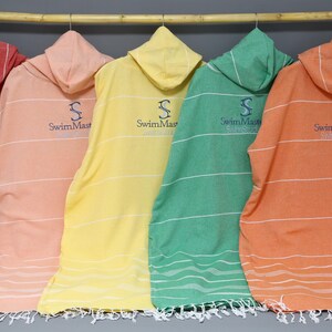 May include: Five hooded towel ponchos in red, light pink, yellow, green, and orange, each featuring thin horizontal white stripes and a dark blue embroidered logo that reads SwimMastery. Each towel has a fringed hem and is hung on a wooden rod.