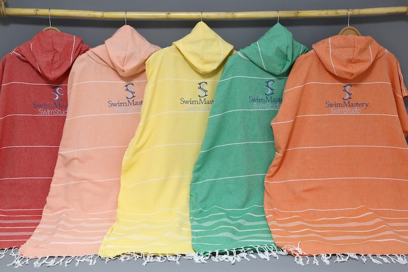 May include: Five hooded towel ponchos in red, light pink, yellow, green, and orange, each featuring thin horizontal white stripes and a dark blue embroidered logo that reads SwimMastery. Each towel has a fringed hem and is hung on a wooden rod.
