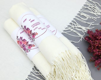 Könnte beinhalten: Drei gerollte cremefarbene Schals mit Fransen. Jeder ist mit einem Etikett mit floralen Akzenten und dem Text "NAME and NAME 12/03/2024 SATURDAY SAVE THE DATE" umwickelt. Die Schals sind auf einer grauen Oberfläche angeordnet.