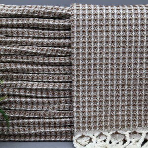 May include: A stack of brown and white waffle weave towels with white fringe. The towels are folded and stacked on a gray surface.