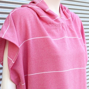 May include: A bright pink hooded poncho featuring a woven texture with thin horizontal white stripes. The garment has short sleeves, a hood, and a straight hemline, displayed on a mannequin to show the drape and the fabric's detailed weave.