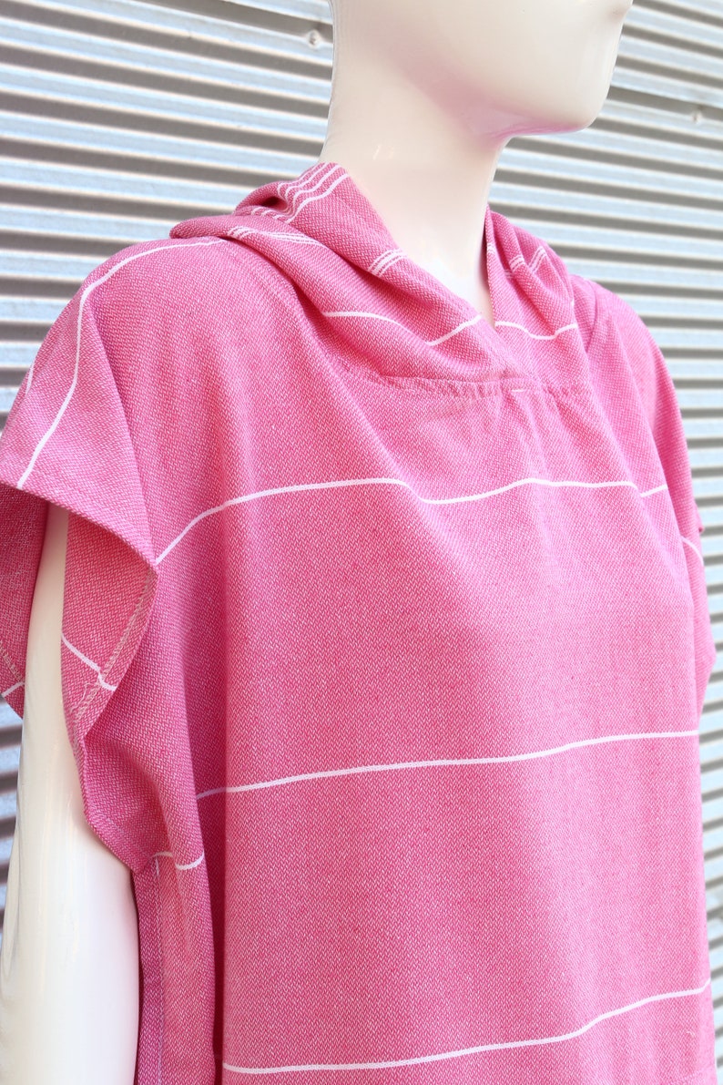 May include: A bright pink hooded poncho featuring a woven texture with thin horizontal white stripes. The garment has short sleeves, a hood, and a straight hemline, displayed on a mannequin to show the drape and the fabric's detailed weave.