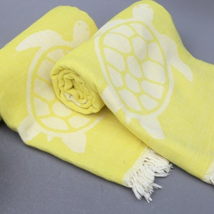Op de afbeelding: Twee gele strandhanddoeken met witte schildpadontwerpen en witte franjes. De handdoeken zijn opgerold en geplaatst op een grijs oppervlak.