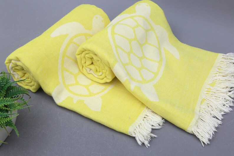 Op de afbeelding: Twee gele strandhanddoeken met witte schildpadontwerpen en witte franjes. De handdoeken zijn opgerold en geplaatst op een grijs oppervlak.