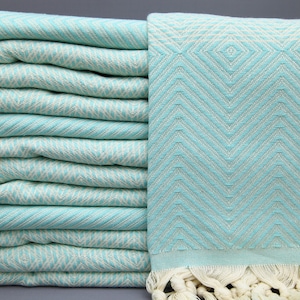 Puede incluir: Una pila de toallas turcas de color azul claro con un patrón de chevron blanco. Las toallas tienen flecos blancos en los extremos.