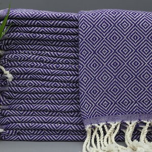 May include: A stack of purple and white patterned Turkish towels with white fringe. The towels are folded and stacked on a gray surface.