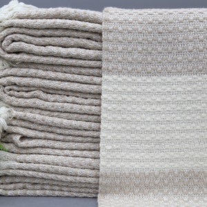 May include: A stack of beige and white woven throw blankets with fringe. The blankets are folded and stacked on a gray surface.
