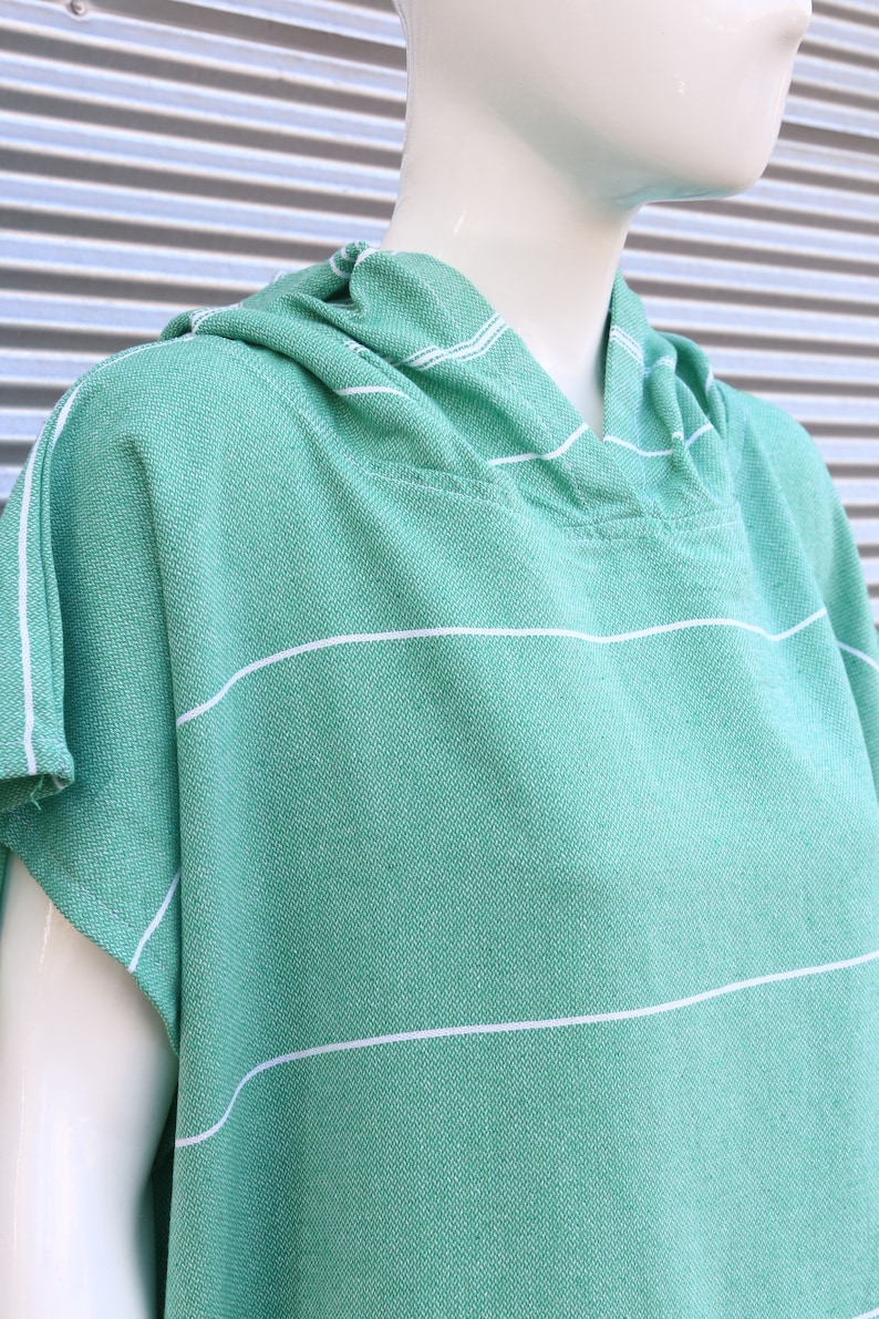 May include: A light green hooded poncho featuring a woven texture and thin horizontal white stripes. The garment has short sleeves and a hood, constructed from a textured fabric with a visible weave pattern, displayed on a white mannequin.