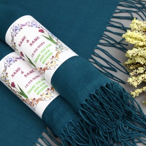 May include: Two teal blue scarves with white labels. The labels have a floral design and the text "NAME & NAME 12/03/2024 THANK YOU FOR JOIN OUR WEDDING".