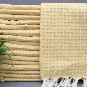May include: A yellow waffle weave Turkish towel with white fringe. The towel is folded and stacked, showing the texture of the fabric.