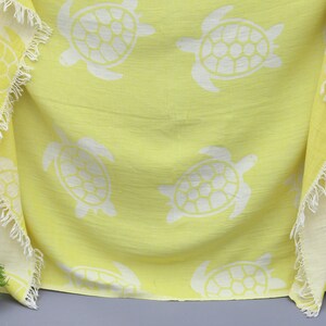 Op de afbeelding: Een gele strandhanddoek met een witte franje rand en een patroon van witte zeeschildpadden.