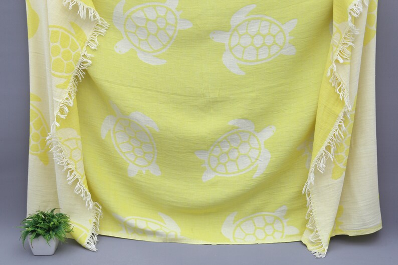 Op de afbeelding: Een gele strandhanddoek met een witte franje rand en een patroon van witte zeeschildpadden.